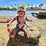 baby, child, crocheted_hat, crocheted_outfit, rock, water, beach, ocean, tropical, palm_trees, sunny, smiling, toy, outdoor, nature, happy, barefoot, infant, blue_sky, vacation