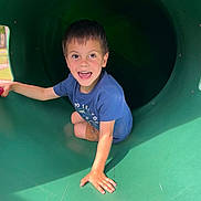 Ashtyn is registered to the contest to win money with this photo: child, boy, playground, slide, smiling, happy, outdoor, tunnel, green, shorts, blue_shirt, sunlight, shadow, play, fun, park, casual_clothing, young, joy, hand