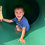 child, boy, playground, slide, smiling, happy, outdoor, tunnel, green, shorts, blue_shirt, sunlight, shadow, play, fun, park, casual_clothing, young, joy, hand