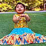 child, toddler, smiling, dress, colorful_clothing, grass, outdoor, happy, clapping, person, nature, greenery, cute, young_child, sitting, portrait, daylight, playful, summer, fun