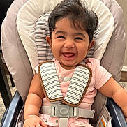 Varadha joined the competition — help win amazing prizes! baby, smiling, high_chair, child, pink_clothing, harness, indoor, happy, cute, seatbelt, infant, person, young_child, face, hair, chubby, sitting, arm, safety, seat