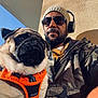 man, dog, pug, sunglasses, beanie, headphones, orange_harness, jacket, camouflage, outdoor, wall, shadow, sunlight, portrait, pet, closeup, casual, facial_hair, human, animal