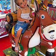 Rose participe au concours pour gagner de l'argent avec cette photo : child, girl, smiling, braided_hair, arcade, mechanical_horse, toy, plush_toys, colorful, carnival, ride, happy, summer_clothing, denim_shorts, sandals, indoor, play, fun, amusement, portrait