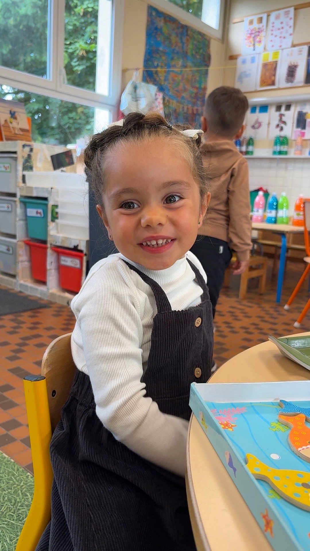 Rose participe au concours pour gagner de l'argent avec cette photo : child, girl, smiling, braided_hair, classroom, table, toy, pinafore_dress, long_sleeve_shirt, chair, floor_tiles, window, backpack, boy, colorful_bottles, artwork, education, indoor, happy, person