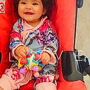 Ariela joined the competition — help win amazing prizes! baby, child, stroller, toy, flower_headband, curly_hair, smiling, red, colorful, clothing, happy, indoor, cute, portrait, sitting, infant, playful, person, accessory, bright