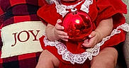 Ariela is registered to the contest to win money with this photo: baby, infant, child, smiling, red_dress, headband, christmas_ornament, plaid_pillow, joy_text, couch, indoors, holiday, festive, white_shoes, lace, happy, sitting, curly_hair, cute, celebration