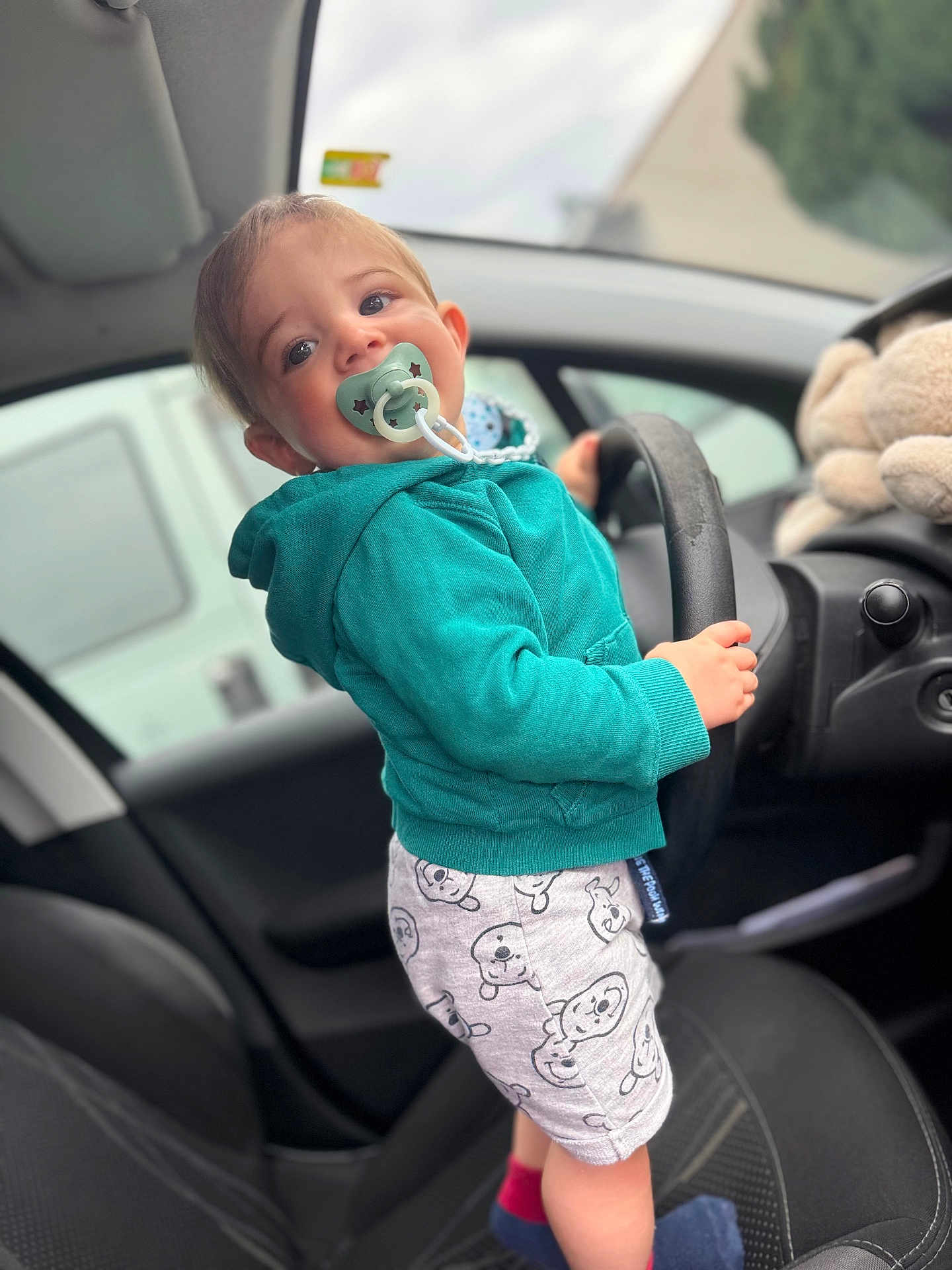 Maylon a rejoint le concours — aidez-le/la à gagner de superbes lots ! toddler, child, car, steering_wheel, pacifier, hoodie, shorts, plush_toy, seat, interior, window, hand, footwear, socks, face, person, young_child, cute, indoor, car_seat