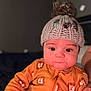 baby, child, knitted_hat, pom_pom, orange_onesie, lion_pattern, face, eyes, hand, indoor, portrait, cute, infant, clothing, person, expression, warm_clothing, young_child, sitting, holding