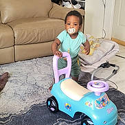 Ahmed participe au concours pour gagner de l'argent avec cette photo : child, toddler, baby, pacifier, pacifier_chain, ride_on_toy, toy_car, standing, living_room, couch, rug, carpet, baby_seat, blanket, floor, playtime, portrait, smiling, furniture, toy_wheels