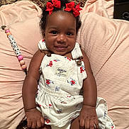 Jadore is registered to the contest to win money with this photo: baby, child, smiling, red_bows, floral_dress, overalls, cushion, blanket, pacifier_clip, beads, indoor, carpet, cute, happy, portrait, infant, clothing, skin, person, lying_down