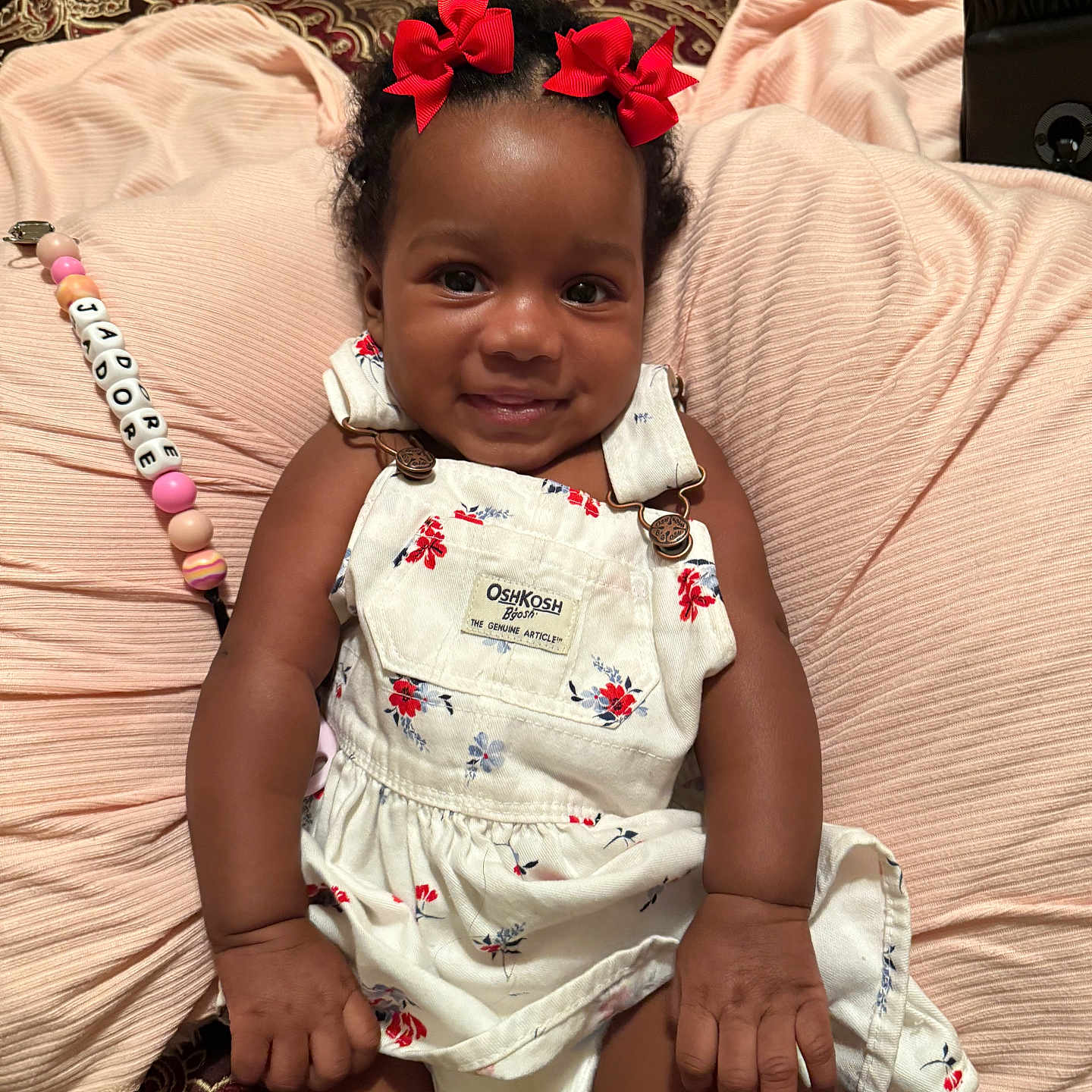 Jadore is registered to the contest to win money with this photo: baby, beads, blanket, carpet, child, clothing, cushion, cute, floral_dress, happy, indoor, infant, lying_down, overalls, pacifier_clip, person, portrait, red_bows, skin, smiling