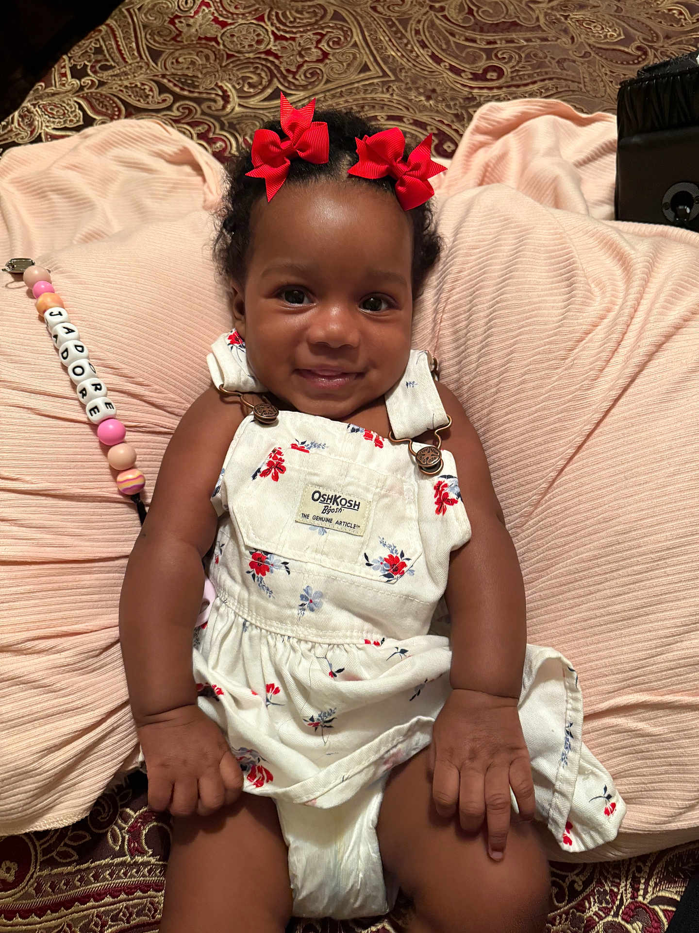Jadore is registered to the contest to win money with this photo: baby, child, smiling, red_bows, floral_dress, overalls, cushion, blanket, pacifier_clip, beads, indoor, carpet, cute, happy, portrait, infant, clothing, skin, person, lying_down