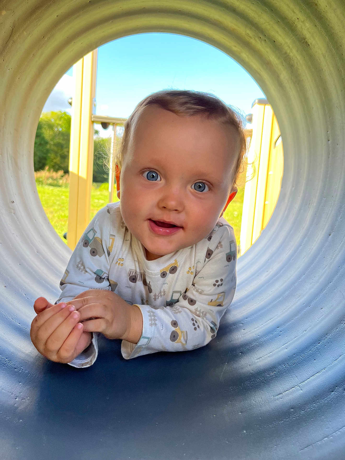 Côme a rejoint le concours — aidez-le/la à gagner de superbes lots ! toddler, child, baby, playground, tunnel, blue_eyes, outdoor, sunny, curious, smiling, long_sleeve_shirt, construction_vehicle_pattern, grass, daylight, cute, portrait, happy, face, head, hands