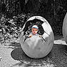 Artūras is registered to the contest to win money with this photo: black_and_white, cap, child, color_pop, cracked_egg, curious, egg_sculpture, face, foliage, gravel, hands, hat, nature, outdoor, peeking, playful, sculpture, shade, statue, toddler