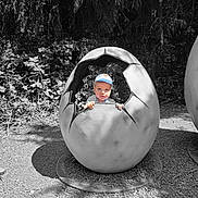 Artūras is registered to the contest to win money with this photo: cap, child, cracked_egg, curious, daylight, egg_sculpture, face, forest, gravel, hat, nature, outdoor, peeking, plants, playful, sculpture, shadows, small_person, summer, toddler