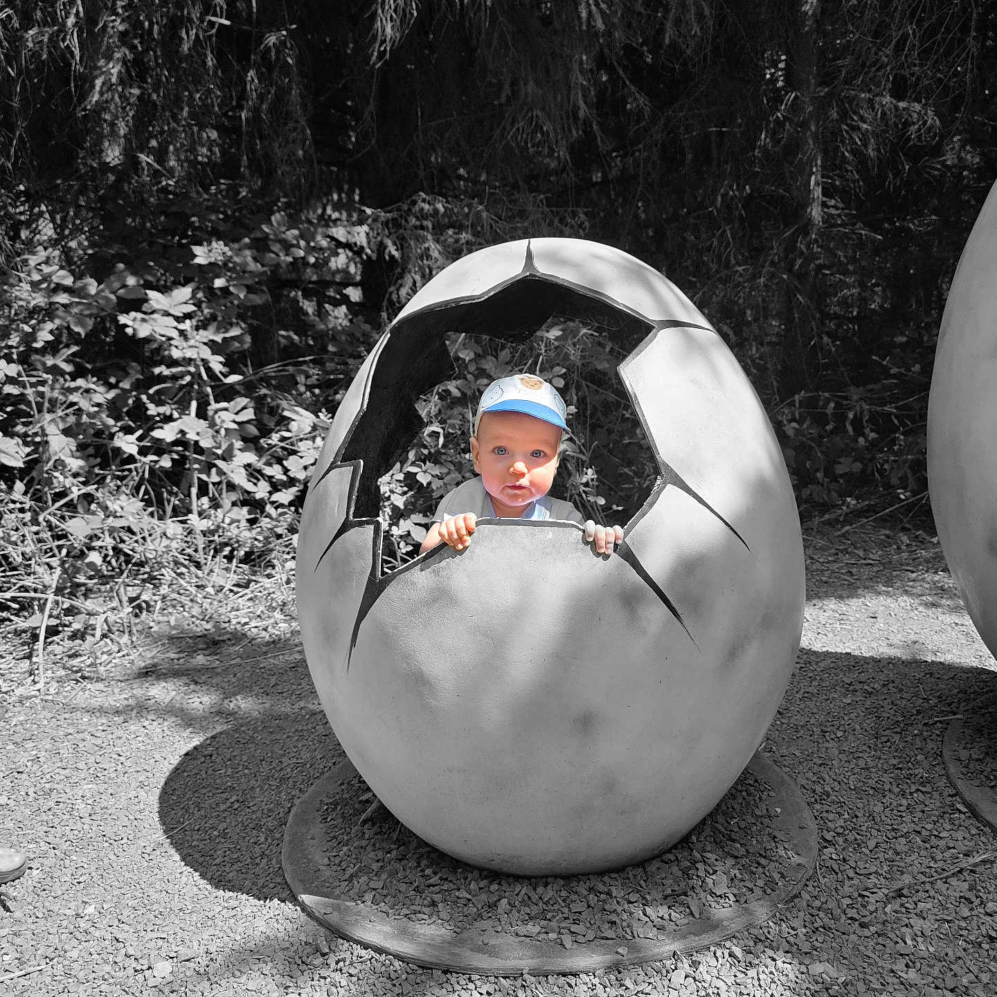 Artūras is registered to the contest to win money with this photo: cap, child, cracked_egg, curious, daylight, egg_sculpture, face, forest, gravel, hat, nature, outdoor, peeking, plants, playful, sculpture, shadows, small_person, summer, toddler