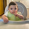 baby, bib, child, curious, cute, eating, face, food, furniture, hand, high_chair, indoor, infant, kitchen, munching, person, snack, table, tile_wall, young_child