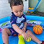 toddler, child, pool, water, toy, basketball, swimsuit, sitting, playing, outdoor, shoes, plastic_pool, yellow_toy, lawn_mower_toy, concrete, fence, summer, water_play, cute, young_child
