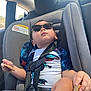 child, toddler, car_seat, sunglasses, cookie, snack, dinosaur_print, seat_belt, car_interior, window, sunlight, casual_clothing, relaxed, seat, sitting, young_child, summer, travel, portrait, cute