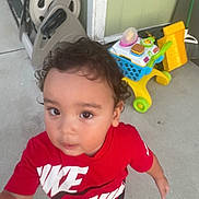 Ambrose is registered to the contest to win money with this photo: child, toddler, curly_hair, red_shirt, nike, toy_shopping_cart, outdoor, concrete, porch, play, casual_clothing, young_child, person, footwear, curious, daylight, sidewalk, home, background_objects, hand