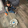 Ambrose joined the competition — help win amazing prizes! toddler, child, floor, mop, toy, chair, jacket, sneakers, wooden_floor, indoor, curly_hair, playful, furniture, home, sitting, black_clothing, blue_pants, cleaning, shadow, casual