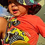 child, toddler, hat, bucket_hat, dinosaur_print, orange_shirt, monster_truck, sippy_cup, outdoor, sunlight, closeup, smiling, face, person, daylight, playful, expression, clothing, headwear, casual