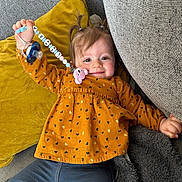 Livia a rejoint le concours — aidez-le/la à gagner de superbes lots ! blanket, blue_pants, child, clothing, couch, cozy, cute, face, happy, home, indoor, orange_top, pacifier, person, pigtails, pillow, relaxing, smiling, toddler, toy