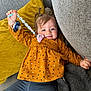 blanket, blue_pants, child, clothing, couch, cozy, cute, face, happy, home, indoor, orange_top, pacifier, person, pigtails, pillow, relaxing, smiling, toddler, toy