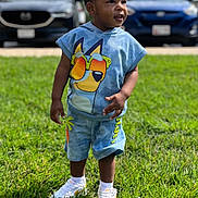 Cairo is registered to the contest to win money with this photo: toddler, child, grass, outdoor, daytime, casual_clothing, shorts, hoodie, sneakers, shoes, person, standing, smiling, cute, young_child, sunlight, parked_cars, background, greenery, happy