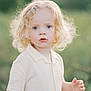 child, toddler, curly_hair, blonde_hair, blue_eyes, cream_shirt, short_sleeves, outdoor, green_background, portrait, innocent_expression, person, young_child, nature, soft_light, summer, standing, curious, closeup, cute