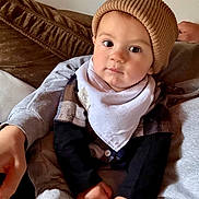 Frederick is registered to the contest to win money with this photo: baby, child, hat, knit_hat, bib, clothing, face, eyes, hand, couch, cushion, person, indoor, sitting, soft_furniture, warm_colors, casual, portrait, infant, cozy