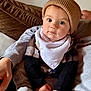 baby, child, hat, knit_hat, bib, clothing, face, eyes, hand, couch, cushion, person, indoor, sitting, soft_furniture, warm_colors, casual, portrait, infant, cozy