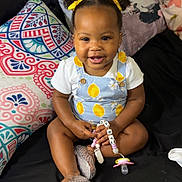 Londyn is registered to the contest to win money with this photo: toddler, child, smiling, sitting, bed, pillow, pacifier, hair_buns, yellow_ribbons, clothing, indoor, cute, happy, person, footwear, shoes, black_bedspread, colorful_pillow, beads, accessory