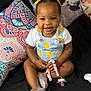 toddler, child, smiling, sitting, bed, pillow, pacifier, hair_buns, yellow_ribbons, clothing, indoor, cute, happy, person, footwear, shoes, black_bedspread, colorful_pillow, beads, accessory