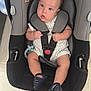 baby, baby_toy, black_gray_car_seat, blue_eyes, car_seat, child, cute, floor_tiles, footwear, indoor, infant, looking_up, navy_sandals, orange_shorts, person, safety_harness, seat, small_hands, striped_clothing, tattoo