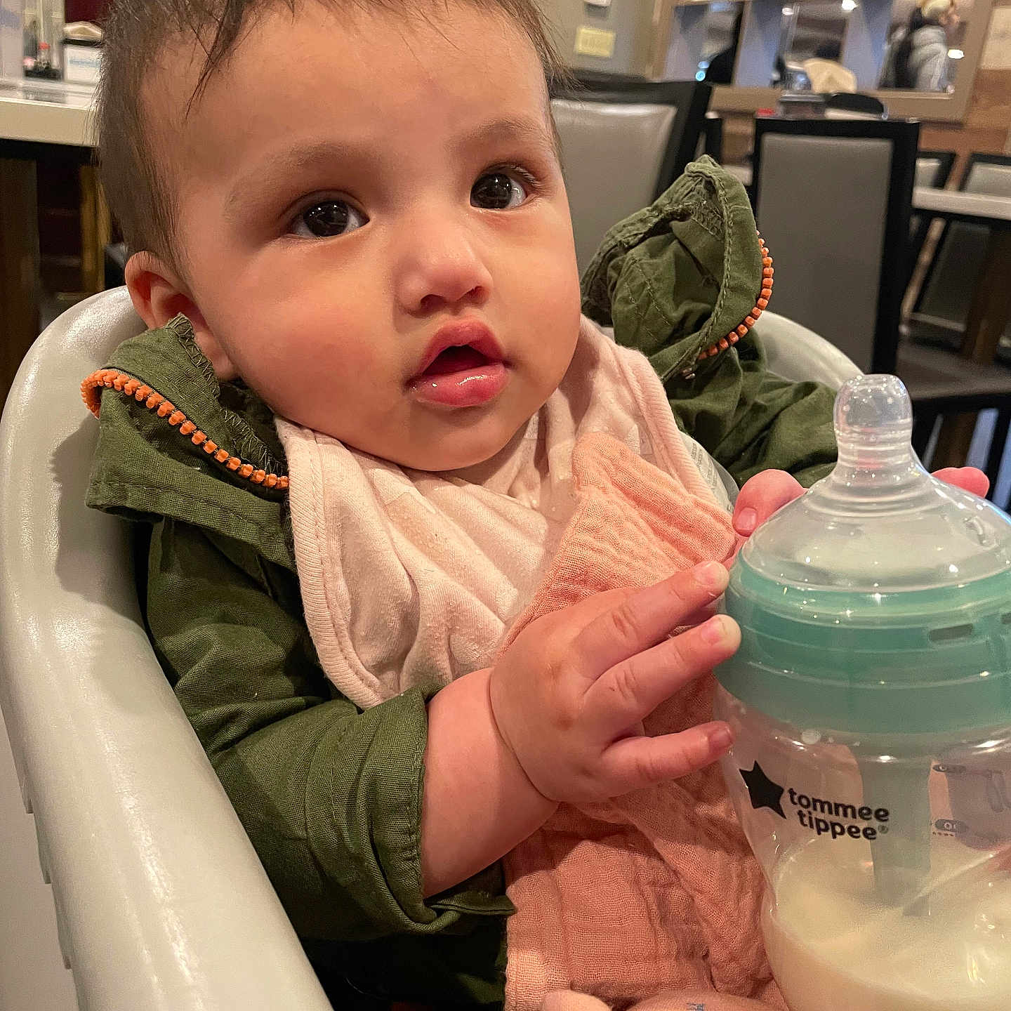 Camila is registered to the contest to win money with this photo: baby, bib, bottle, chair, child, cute, dining_room, expression, face, feeding, green, hand, high_chair, indoor, infant, jacket, milk, person, pink, table