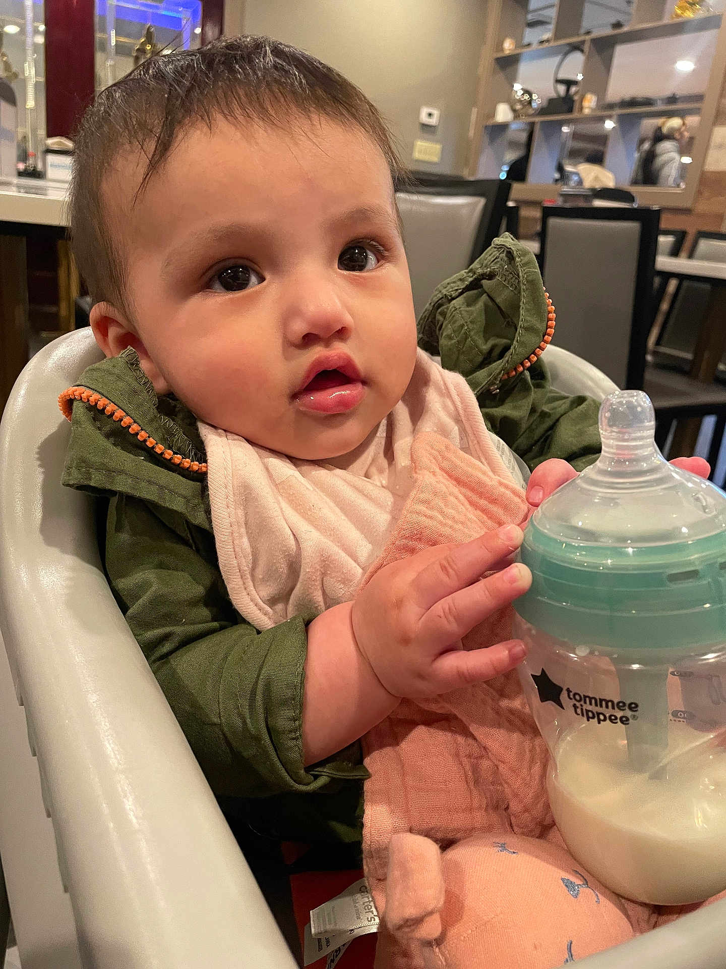 Camila is registered to the contest to win money with this photo: baby, bottle, high_chair, indoor, child, milk, jacket, bib, pink, green, face, hand, table, chair, dining_room, person, cute, infant, feeding, expression