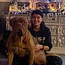 brown_dog, cabinet, carpet, casual_clothing, cozy, decor, dog, festive, furniture, holiday_decorations, home, indoor, person, pet, portrait, santa_figurine, short_hair, sitting, smiling, string_lights