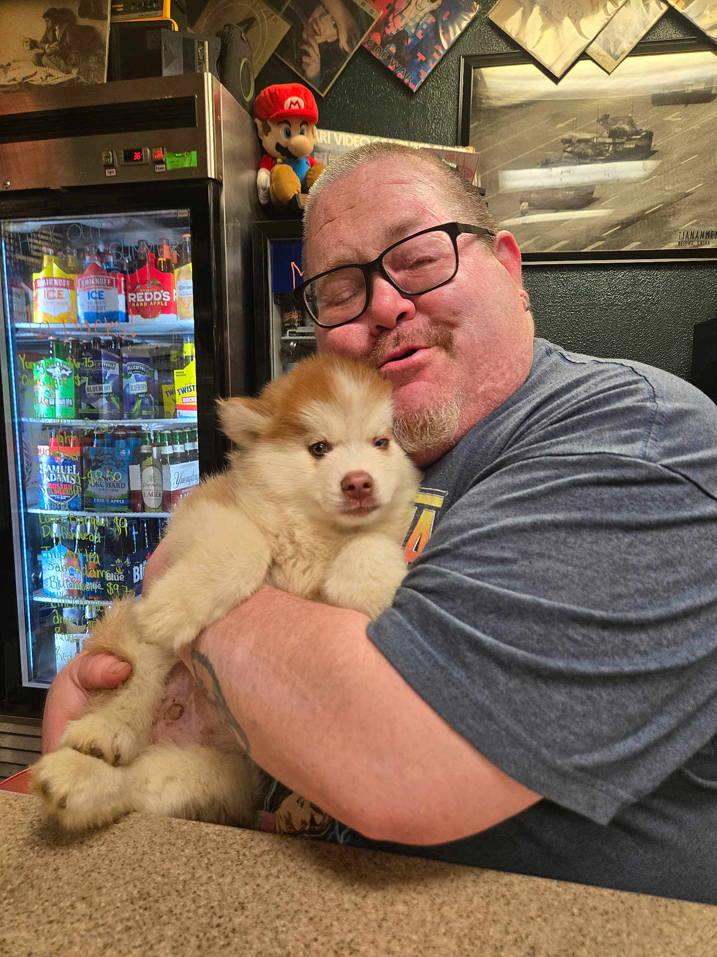 Emmitt is registered to the contest to win money with this photo: man, glasses, puppy, dog, hugging, countertop, refrigerator, beverage, plush_toy, mario, tattoo, casual_clothing, indoor, smiling, portrait, cute, pet, happy, animal, friendly