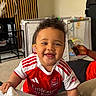 baby, child, smiling, arsenal_jersey, red_and_white_clothing, sock, indoor, living_room, playpen, television, person, furniture, couch, phone, hand, curly_hair, carpet, toy, happy, portrait