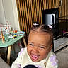 Gia Elora is registered to the contest to win money with this photo: toddler, child, smiling, laughing, indoor, toy, wooden_toy, table, hair_clips, ponytails, sweater, cozy, carpet, furniture, television, modern, person, happy, cute, playful