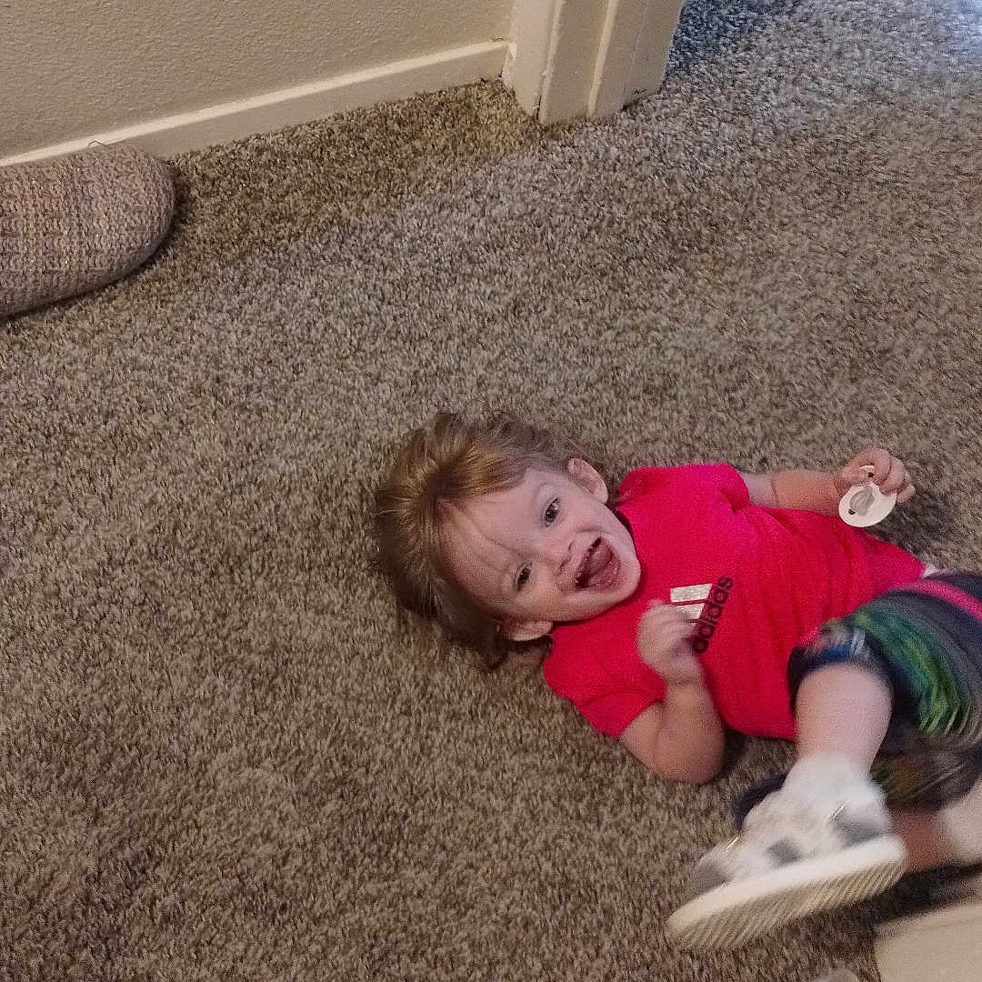 Aspen is registered to the contest to win money with this photo: baby_bottle, candid, carpet, child, colorful_pants, doorway, floor, footwear, happy, home, indoors, pacifier, person, playful, red_shirt, slipper, smiling, toddler, toy, young_child