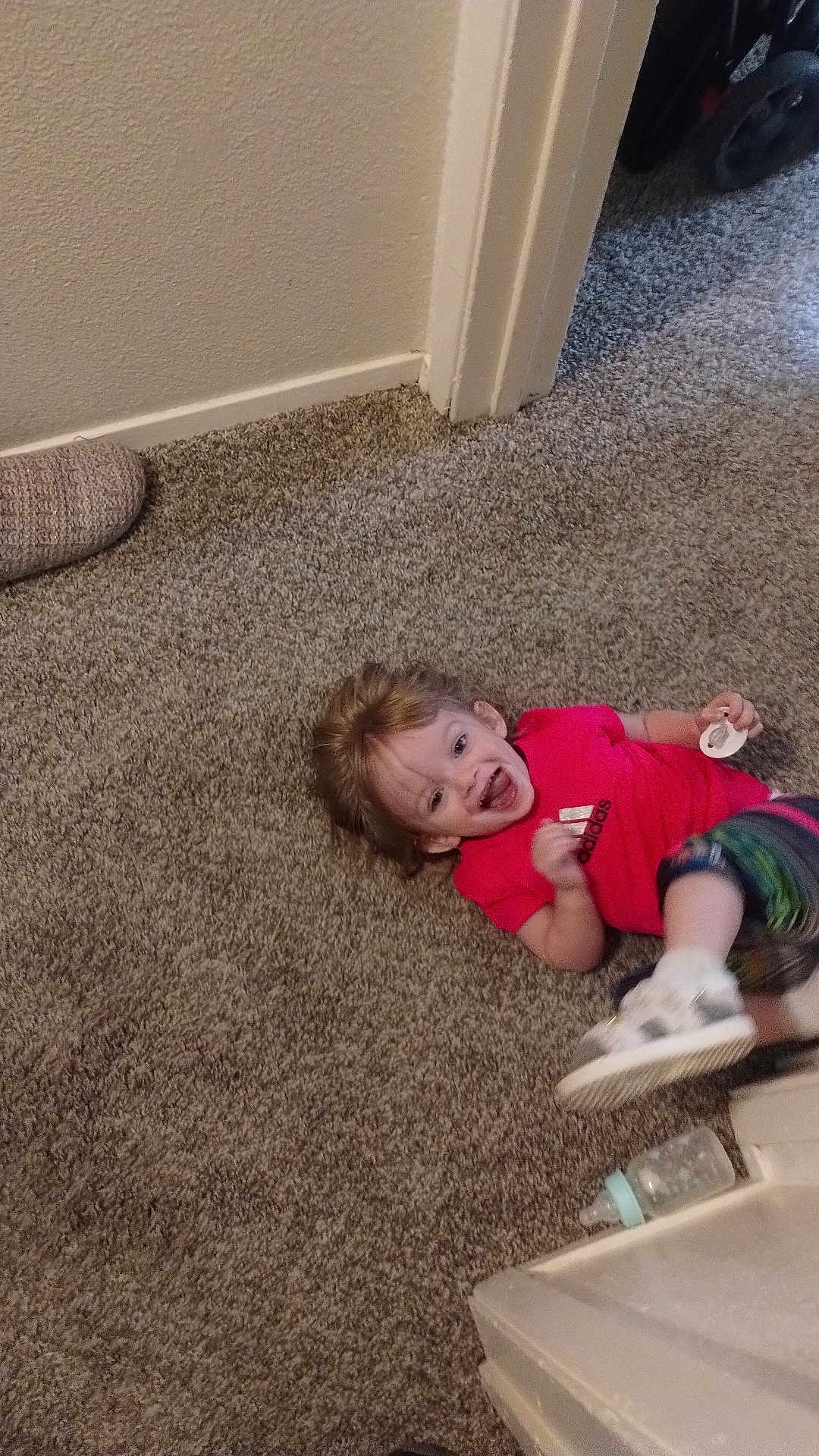 Aspen is registered to the contest to win money with this photo: toddler, child, carpet, floor, doorway, pacifier, baby_bottle, slipper, red_shirt, colorful_pants, smiling, happy, indoors, person, footwear, toy, candid, playful, young_child, home