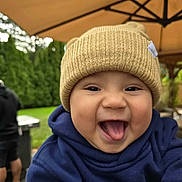 Noah is registered to the contest to win money with this photo: baby, child, hat, knit_hat, hoodie, tongue_out, smiling, outdoor, umbrella, greenery, happy, cute, infant, face, person, apparel, headwear, playful, closeup, daytime