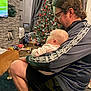 baby, casual_clothing, child, christmas_tree, curtains, dog, family, furniture, glasses, holiday_decorations, indoor, living_room, man, ornaments, person, photo_frame, seated, sports, stone_wall, television