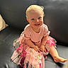 toddler, child, smile, pink_dress, curly_hair, blue_eyes, sitting, black_couch, indoor, natural_light, happy, cute, baby, foot, hand, portrait, clothing, window, skin, young