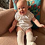 baby, infant, smiling, sitting, floral_pillow, cushion, armchair, white_outfit, knee_socks, bracelet, chubby_legs, cute, portrait, indoor, soft_texture, nursery_decor, wallpaper, happy, legwear, pillowcase
