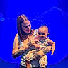 woman, baby, tattoos, smile, holding, floral_outfit, blue_background, jellyfish, aquarium, person, child, portrait, casual_clothing, happy, indoor, arm, head, face, cute, love