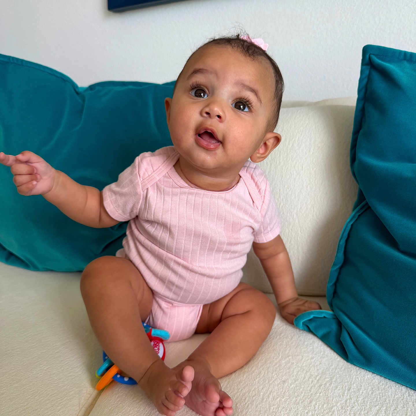 Katalina is registered to the contest to win money with this photo: baby, child, infant, pink_onesie, couch, sofa, teal_cushions, pointing, curious, indoors, expression, face, person, sitting, barefoot, home, furniture, cute, portrait, young