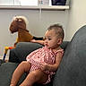 baby, child, striped_clothing, couch, indoor, plush_toy, rocking_horse, furniture, curly_hair, person, toddler, relaxed, seated, soft_toys, home_interior, casual, infant, cute, living_room, young_child